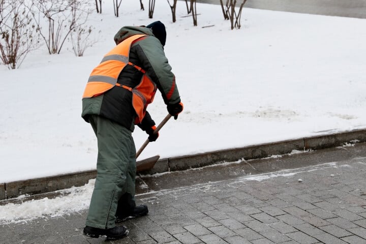 Worker in uniform with a shovel clears the snow off a sidewalk